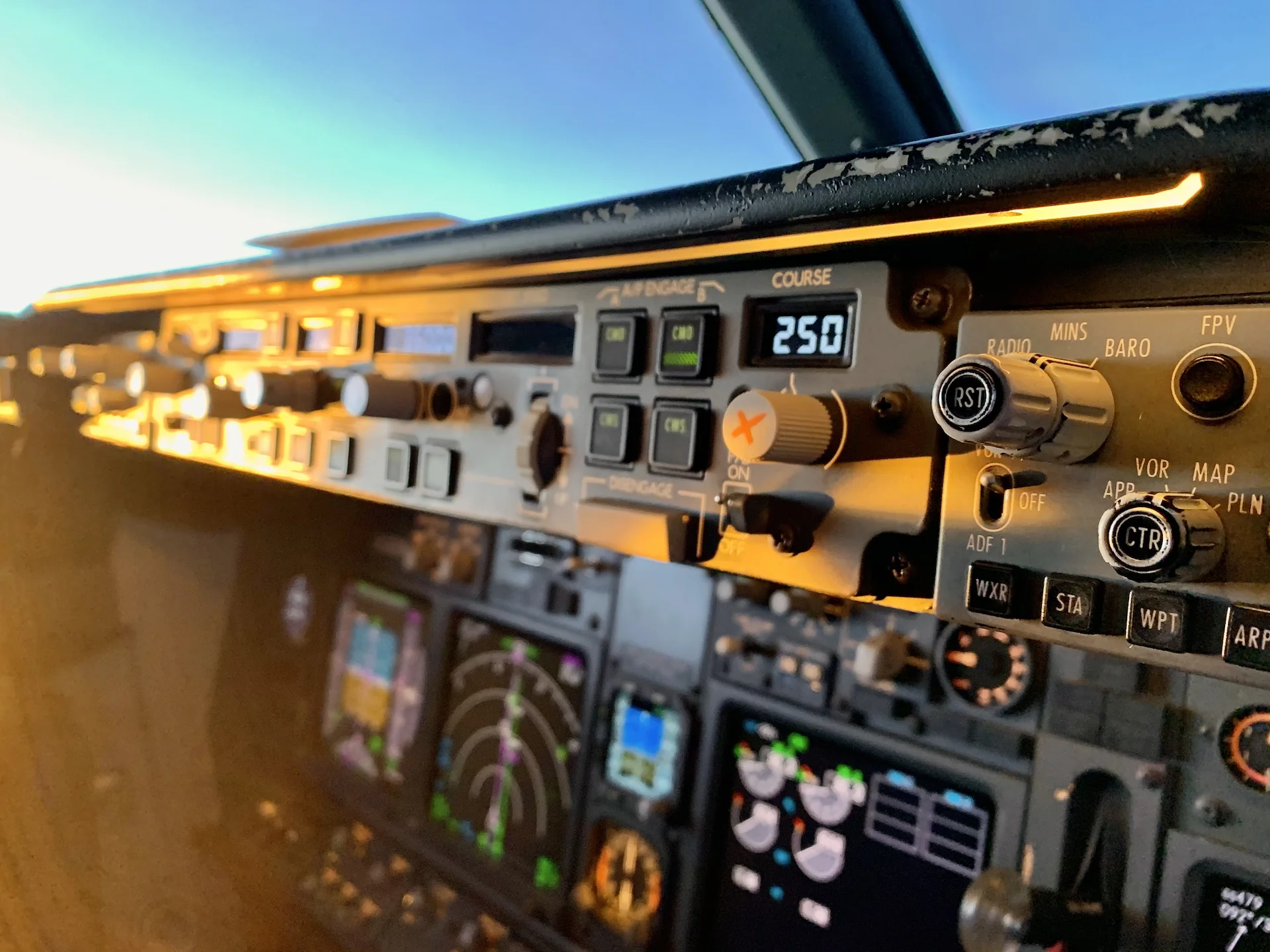 Close-up view of an airplane cockpit control panel showing knobs, buttons, and digital displays, with flight instruments and navigation screens visible in the background.