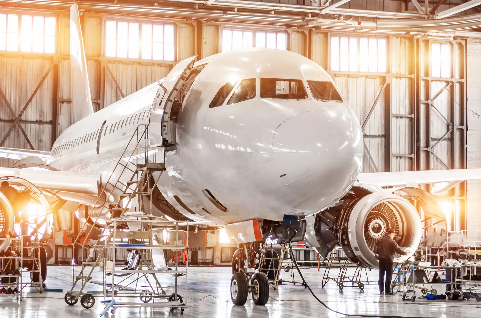 A mechanic inspects and works on a jet engine of an airplane inside a brightly lit aircraft hangar, surrounded by tools and equipment.