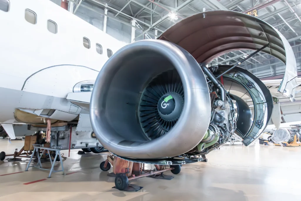 A close-up view of a jet engine being serviced on a passenger airplane inside a brightly lit hangar, with engine panels open to reveal internal components.