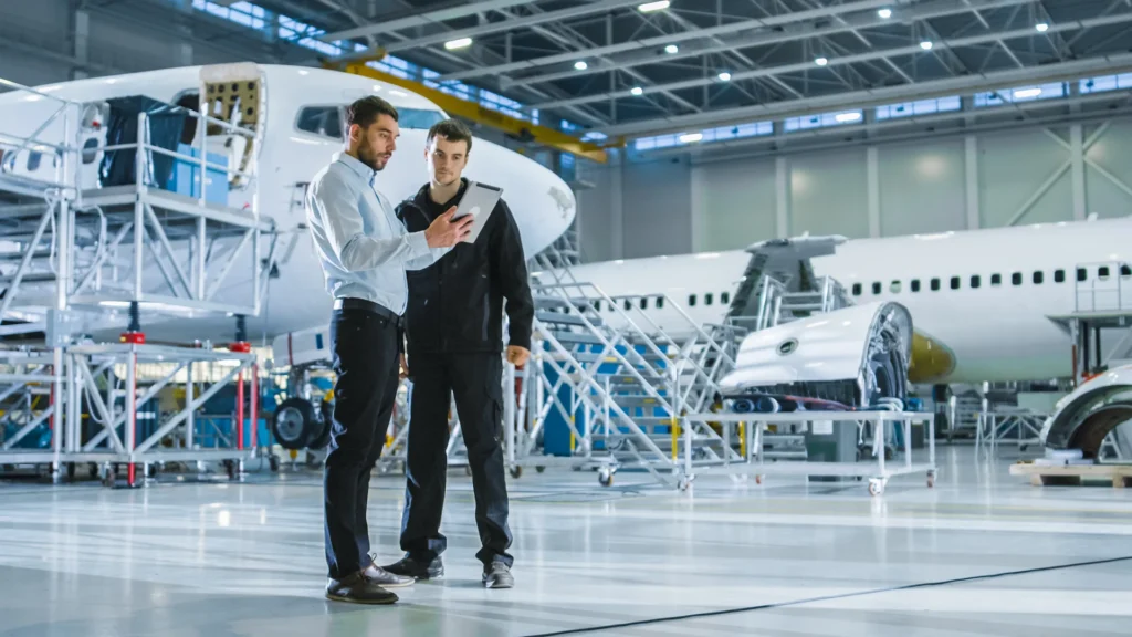 Two men stand in an aircraft hangar, looking at a tablet. One wears a white shirt and dark pants, the other is in a black work uniform. Airplane parts and a jet engine are visible in the background.