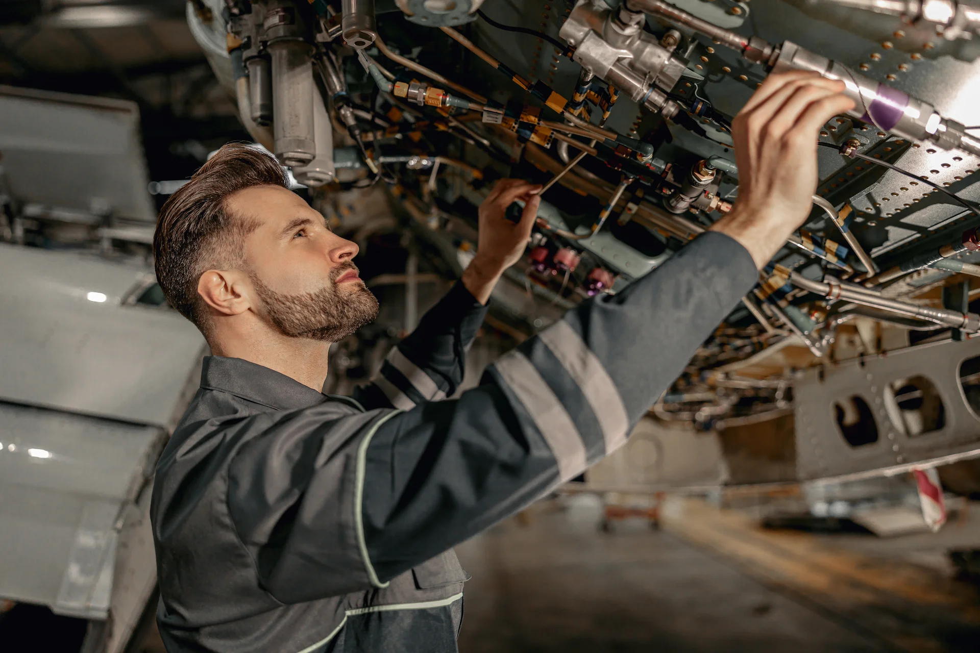 A technician inspects and works on mechanical components under an aircraft, focused on intricate tubing and equipment.