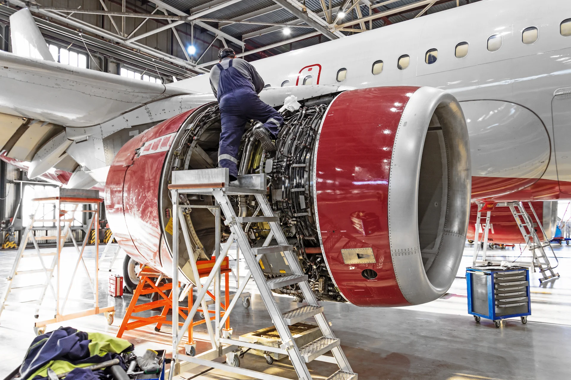 A technician works on the engine of an aircraft inside a hangar, using a ladder and surrounded by tools and equipment.