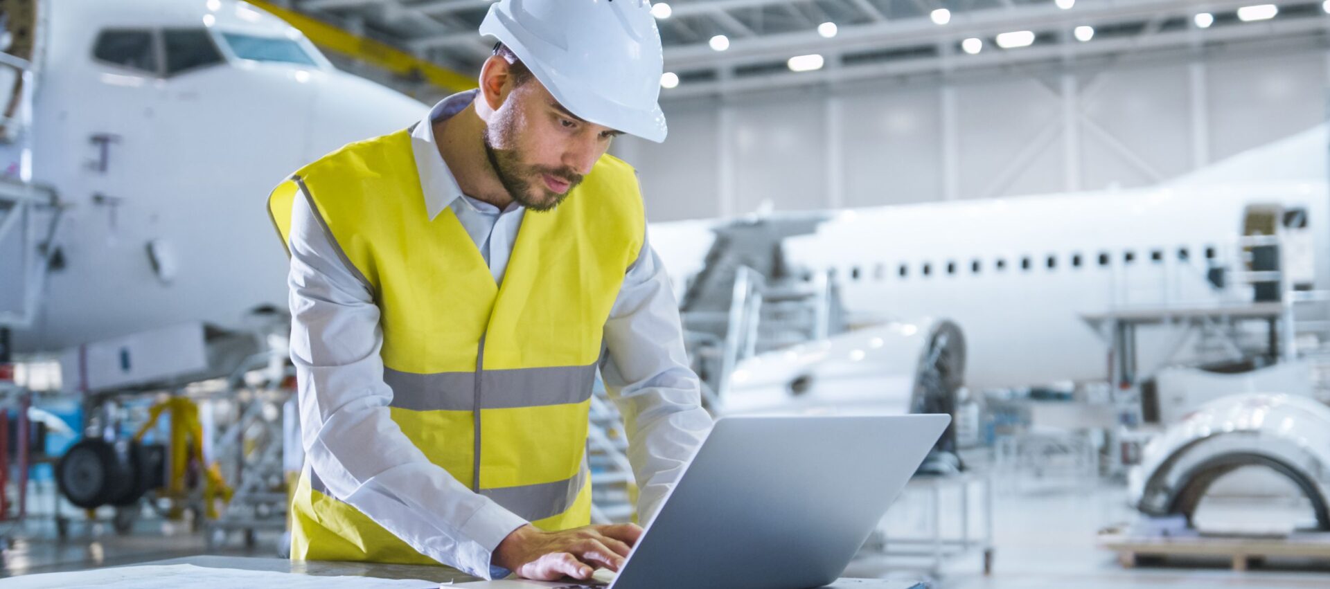 A man in a yellow safety vest and hard hat working on a laptop in an aircraft hangar, with an airplane in the background.