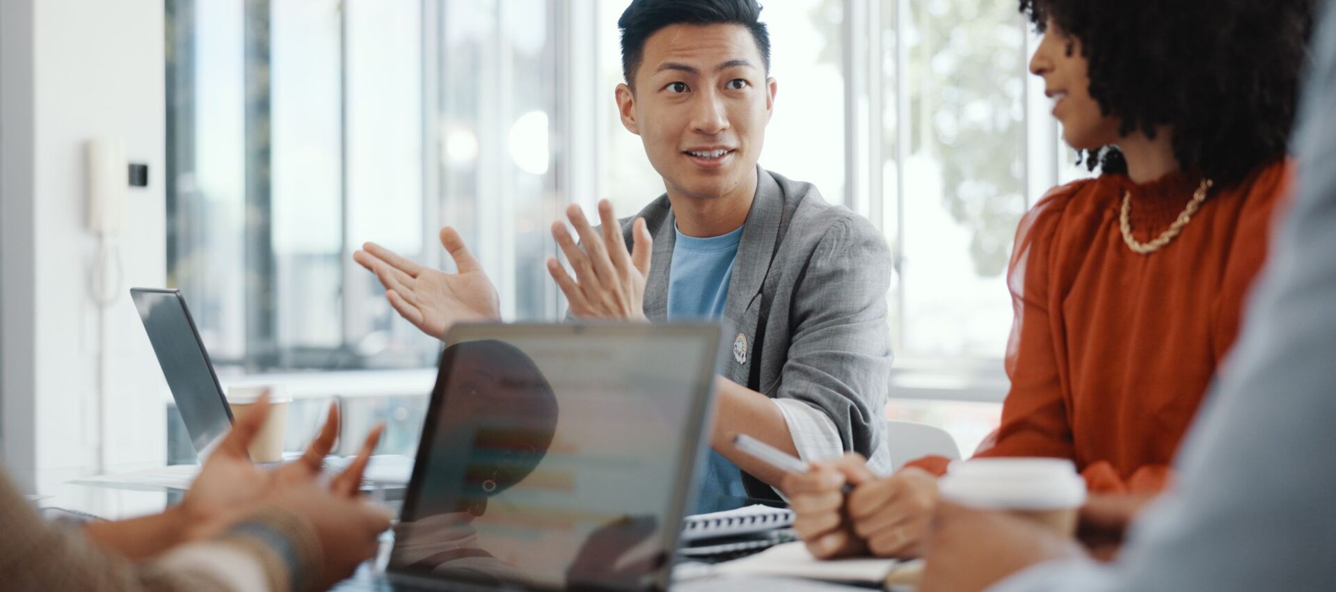 A diverse group of people in a meeting, with one person animatedly speaking and gesturing. Laptops and notebooks on the table.