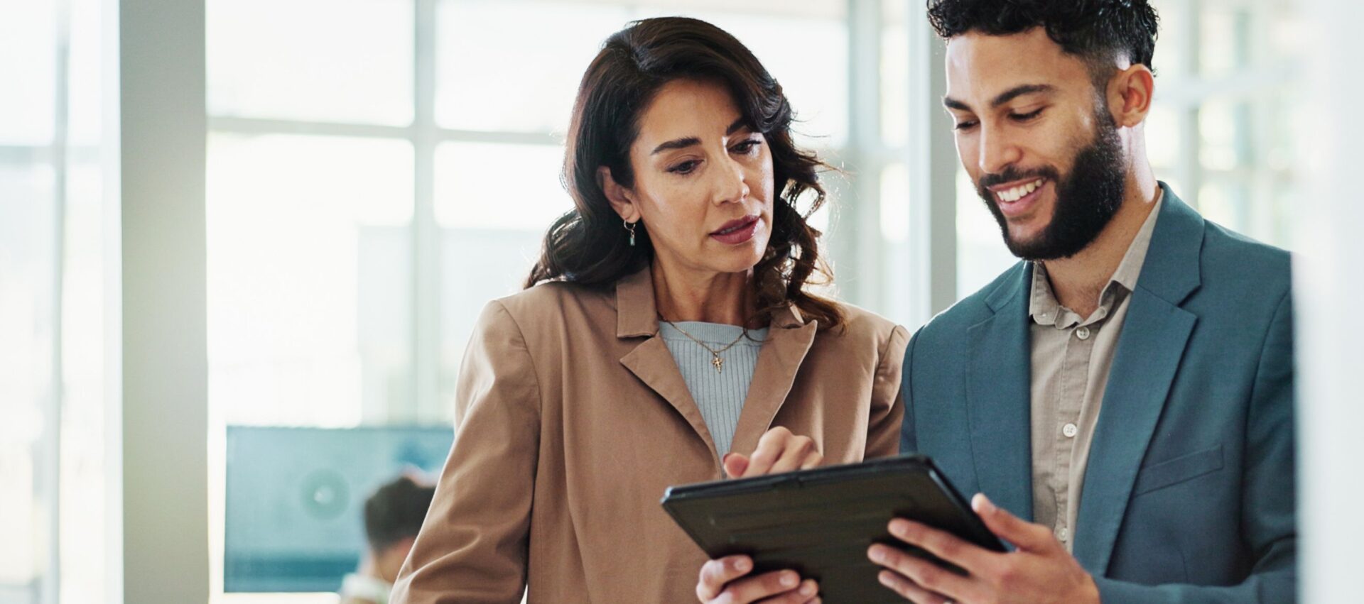 A woman and a man discuss while looking at a tablet in a bright, modern office setting.