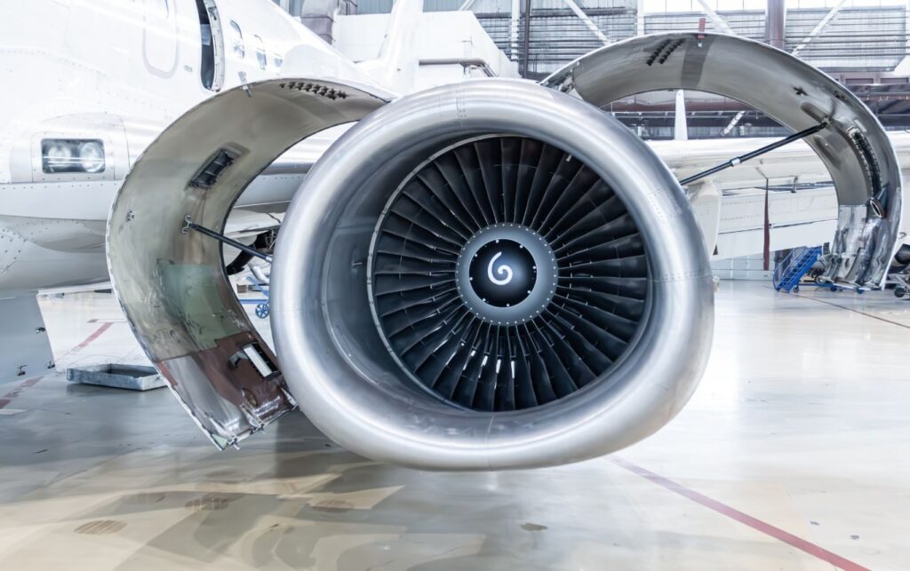Close-up of a jet engine mounted on an aircraft, showcasing the turbine and a shiny silver casing in a hangar setting.