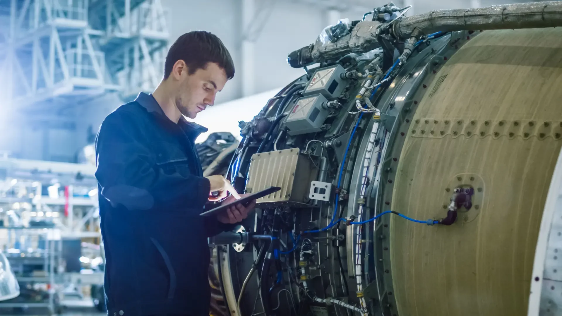 A technician examines an aircraft engine with a tablet in a workshop, displaying intricate machinery and blue wiring.