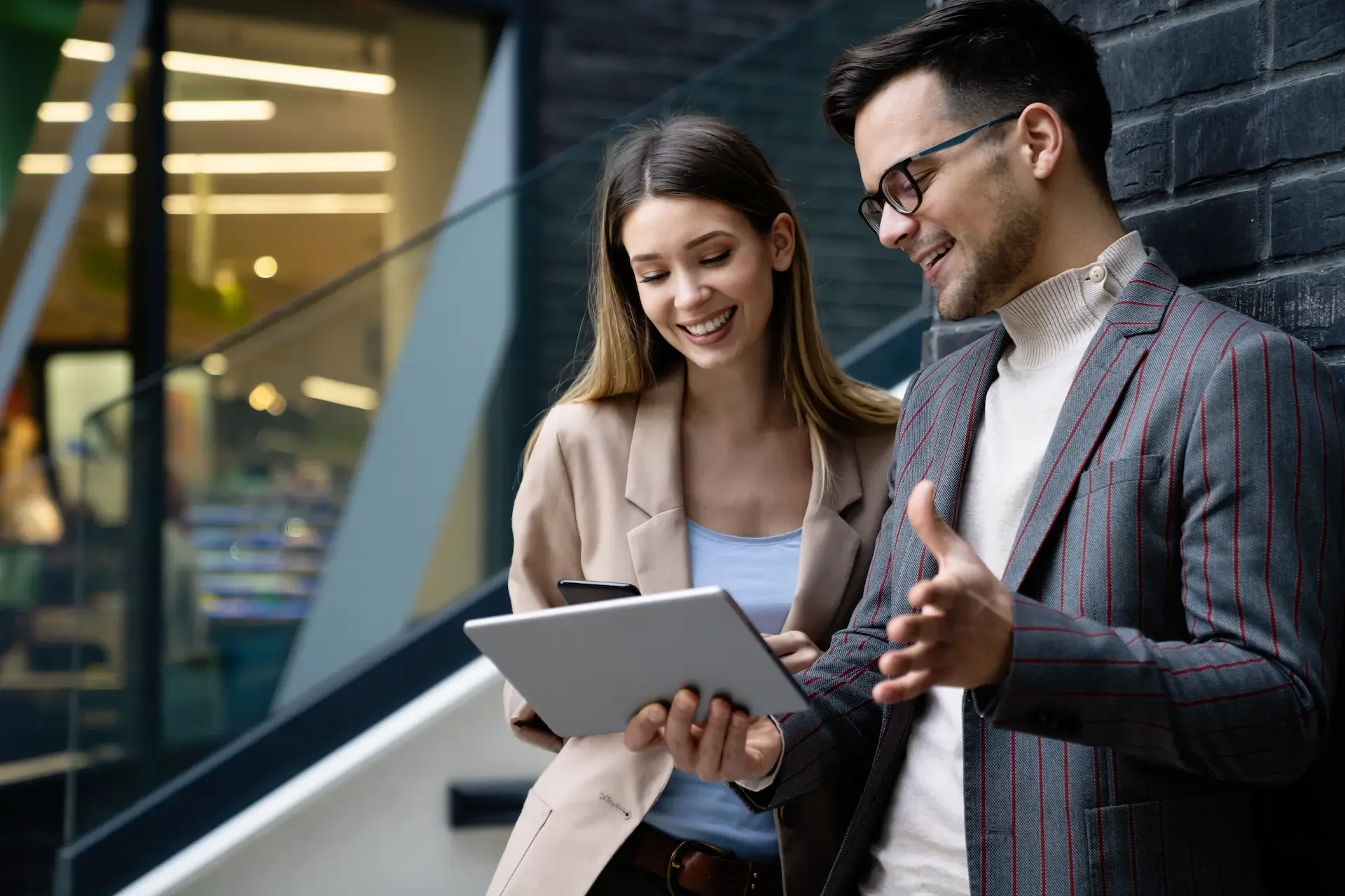 A woman and a man are smiling and discussing while holding a tablet and a smartphone in a modern, stylish setting.